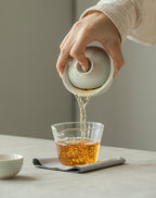 Person pouring tea from a white teapot into a glass cup on a neutral background