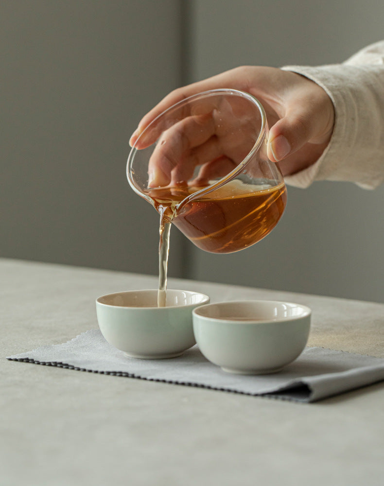 Person pouring tea from a glass teapot into two white ceramic cups on a light surface.