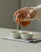 Person pouring tea from a glass teapot into two white ceramic cups on a light surface.